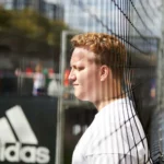 A person with short, light hair stands beside a fence on a sports field, wearing a white T-shirt, possibly taking a break from walking football. In the background, there's a blurred view of tall buildings and a black Adidas sign. The sky above is partly cloudy. © Powerleague